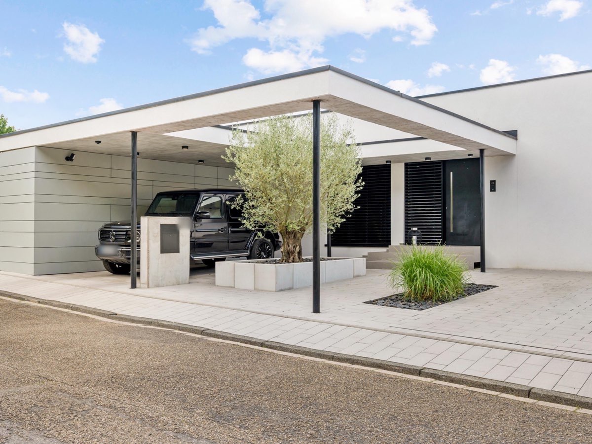 Bungalow mit Flachdach-Carport auf Stahlstützen, Olivenbaum im Betontrog und heller Pflasterfläche