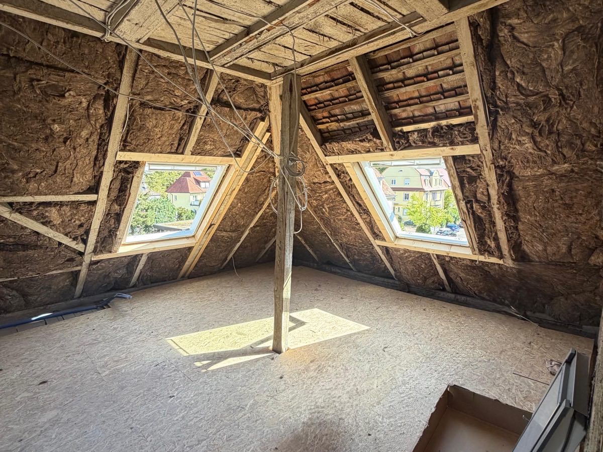 Dachgeschoss im Ausbau: Dämmung zwischen den Sparren, zwei neue Dachfenster mit Blick auf die Nachbarschaft, Holzstütze in der Raummitte, verlegter Boden aus Holzwerkstoffplatten