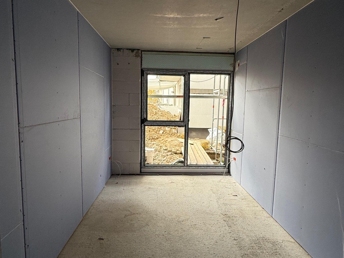 Raum mit fertig beplankten Vorsatzschalen aus blauen Gipsplatten auf beiden Seiten, Leerrohre und Unterputzdosen gesetzt, Estrich eingebracht, Fenster zur Baustelle