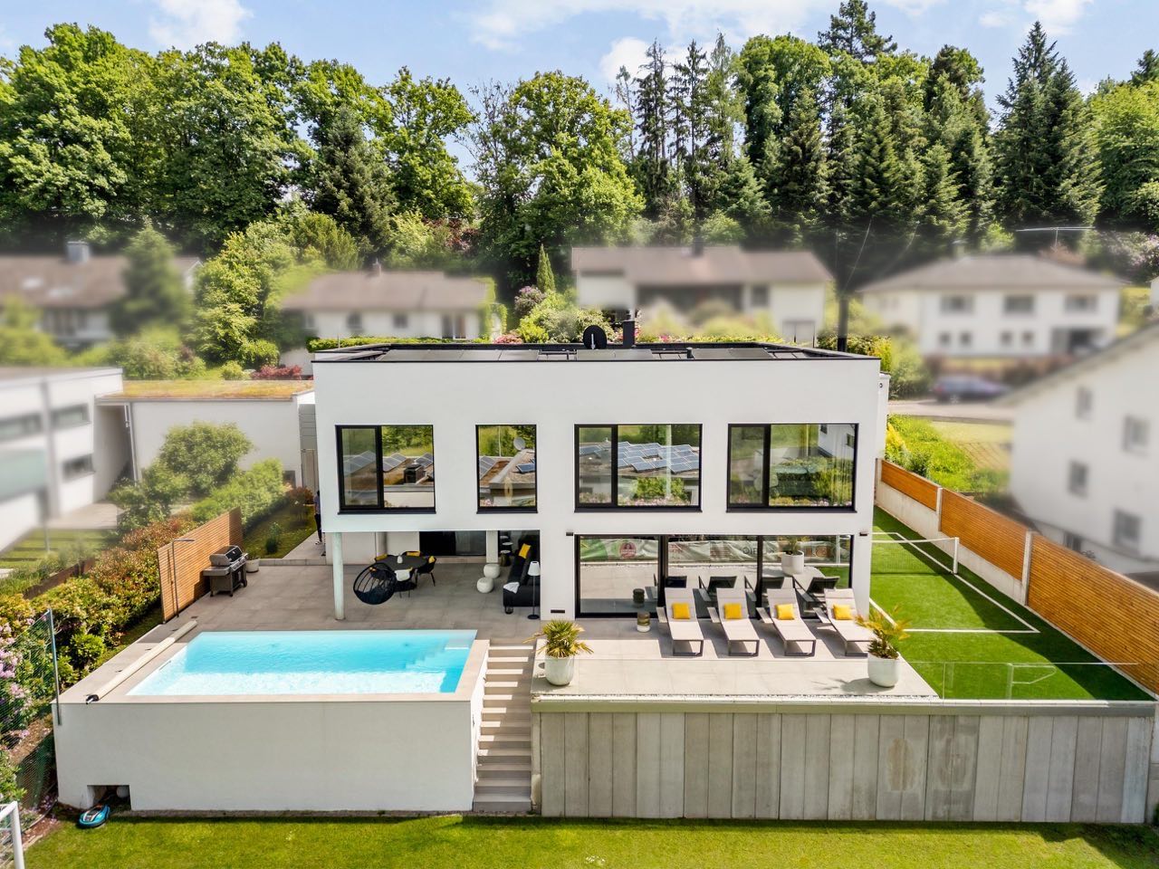 Fertiggestelltes Wohnhaus von MaDe Bauservice aus der Luft: Flachdach mit Photovoltaik, weiße Putzfassade, überdachte Terrasse, Pool, Stützmauer und Rasenfläche
